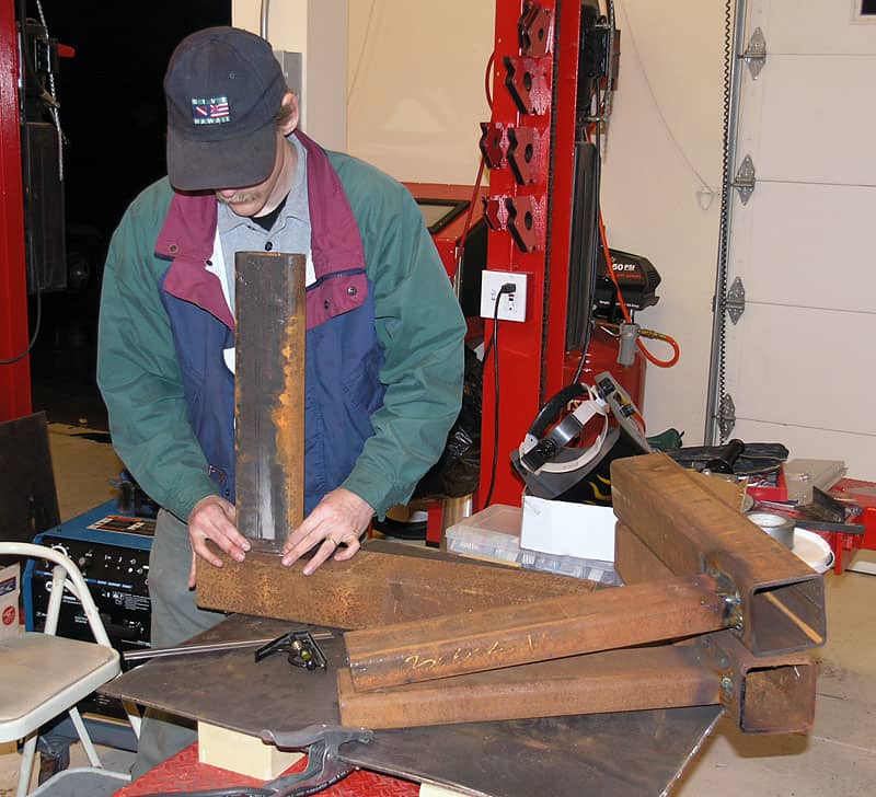A man in a garage workshop examines large metal pieces on a table.