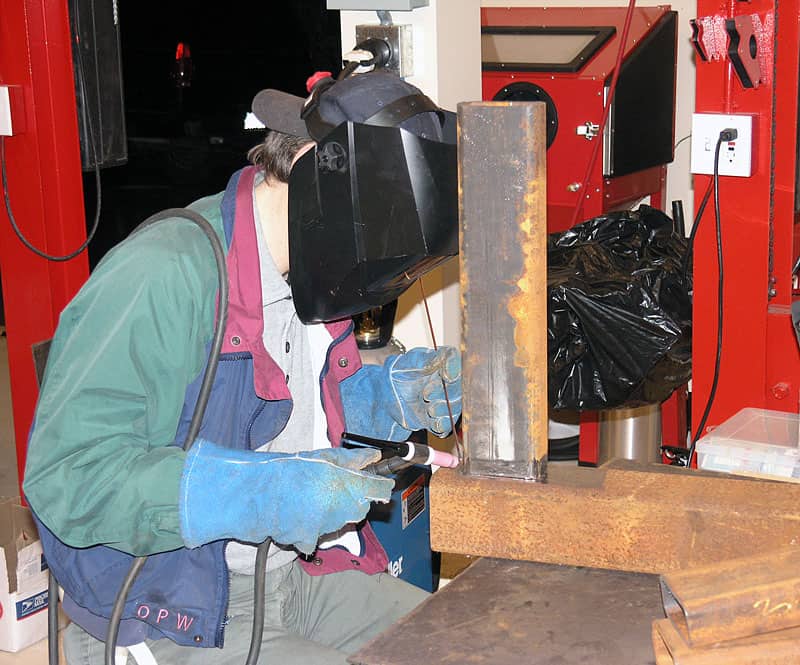 A person in a workshop setting wears protective gear, including a welding helmet, gloves, and jacket.