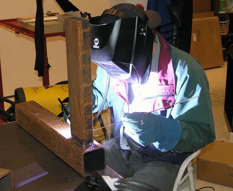 A person wearing a welding helmet and gloves uses an arc welder to join two metal pieces.