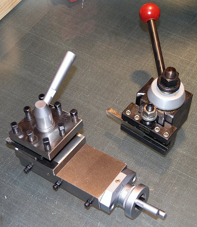 Two metal lathe tool holders on a workbench, one equipped with a lever and the other with a red ball handle, both part of a Quick Change Tool Post system.