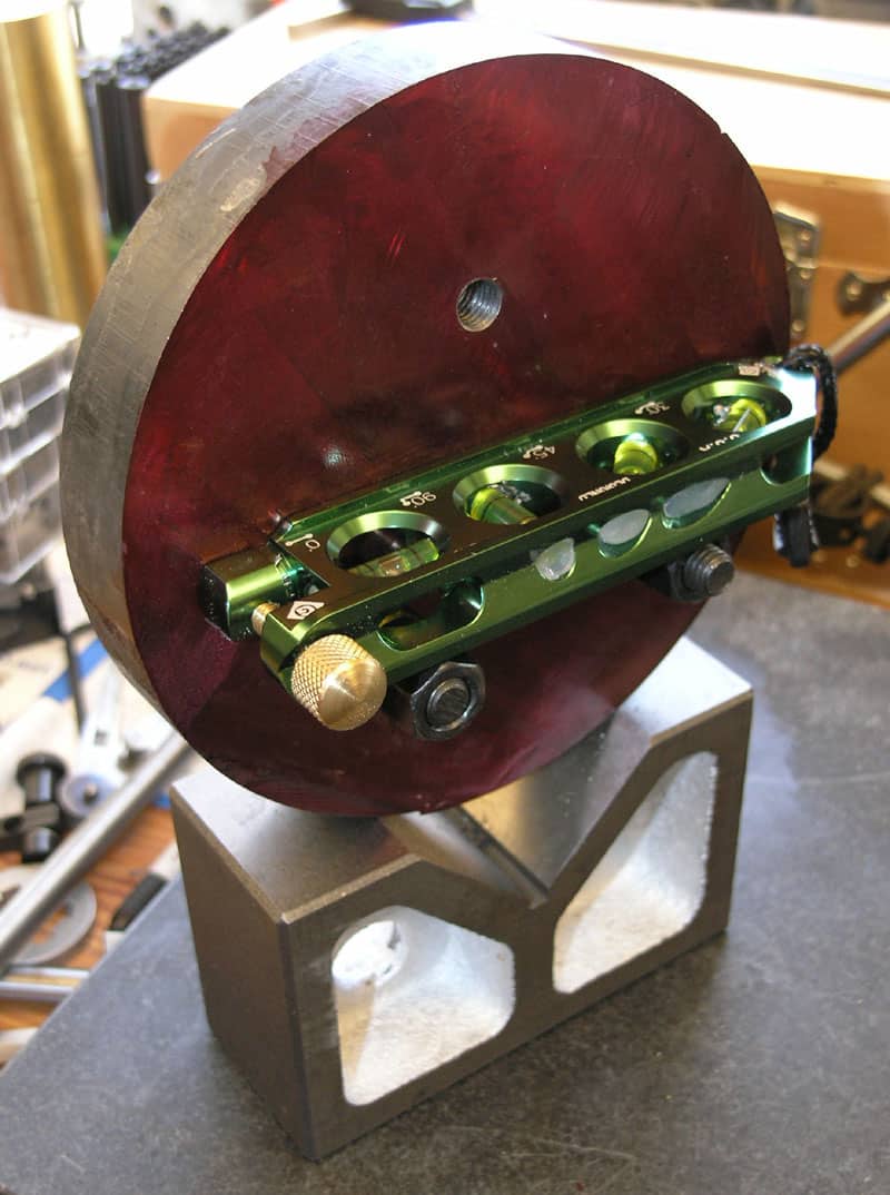 Precision instrument with green metal bar and circular holes, on a red base, with machine shop workshop background.