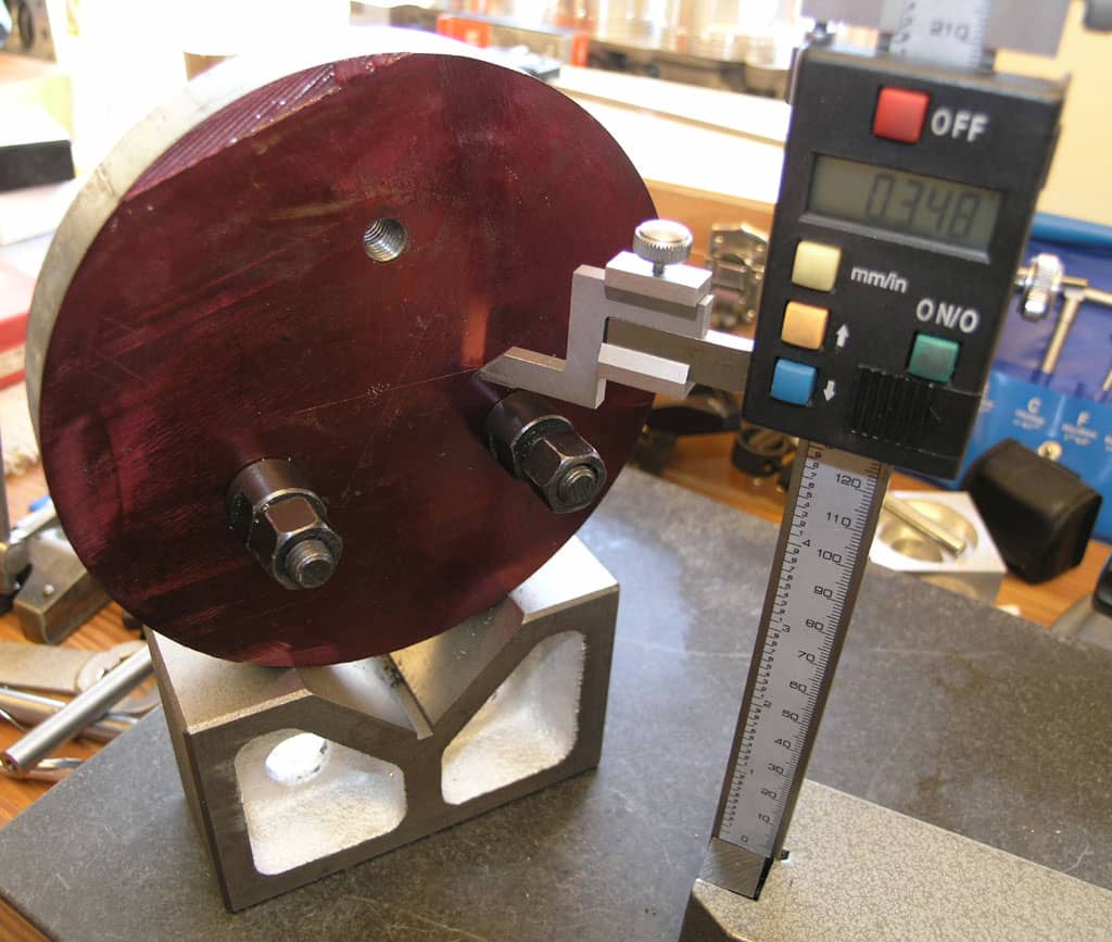 Digital caliper on a metal stand with a red circular object featuring three bolts in a workshop setting.