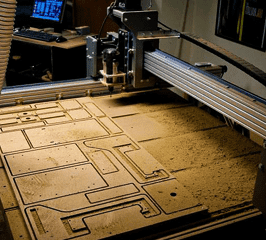 A cnc router in operation, cutting intricate patterns on a wooden board in a workshop with computer monitors visible in the background.