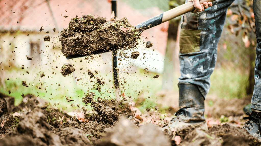 A person in muddy jeans using a shovel to toss soil with an efficient material removal rate, capturing dirt particles mid-air in a vibrant, outdoor setting.