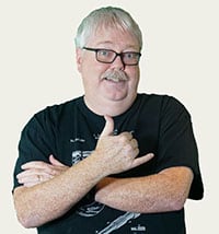 A middle-aged man with short white hair and glasses, wearing a black t-shirt, arms crossed.