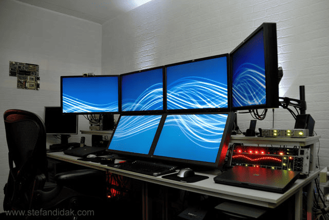 White desk with 4 computer monitors and office supplies against a white brick wall.